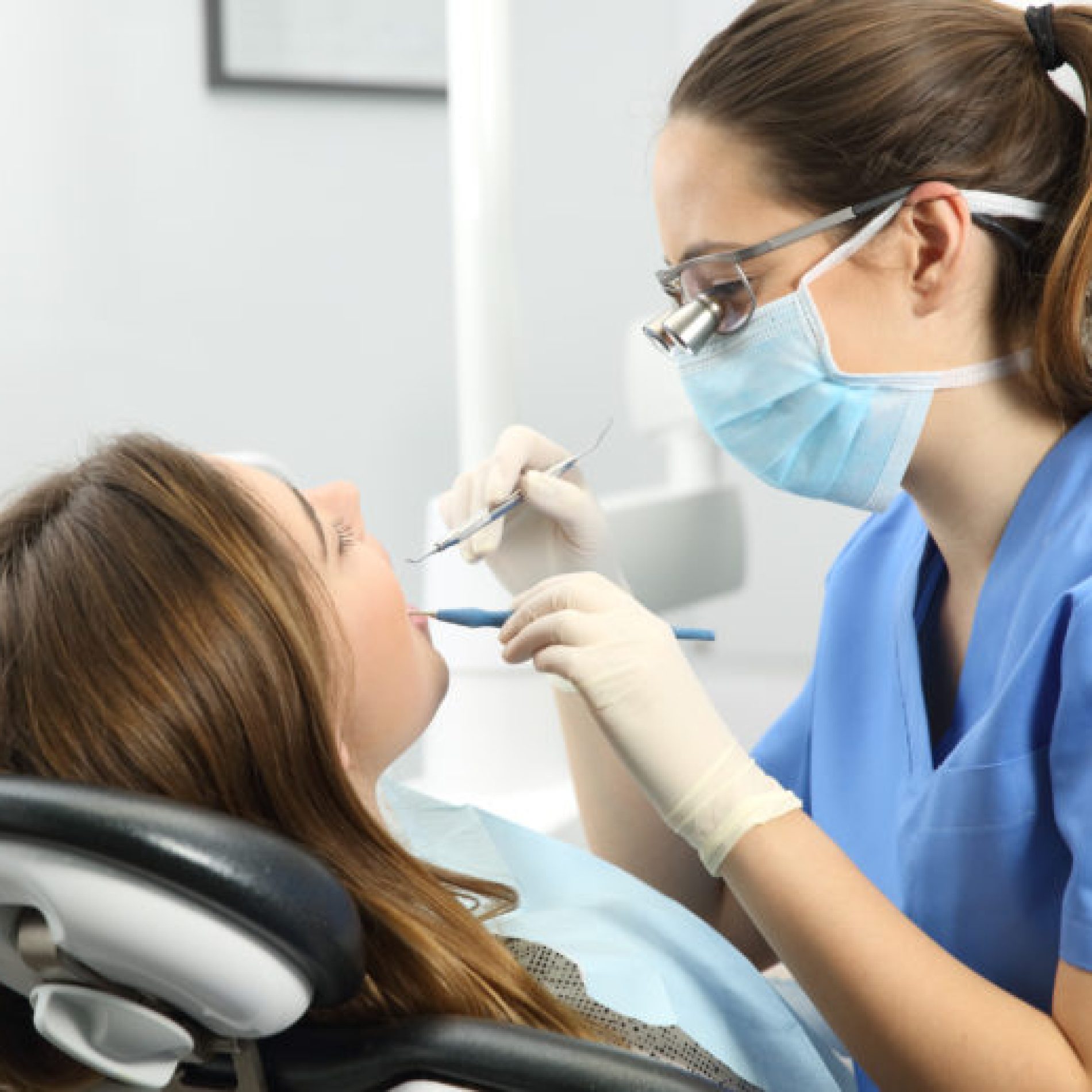 Dentist wearing eyeglasses gloves and mask examining a patient teeth with a dental probe and a mirror in a clinic box with equipment in the background